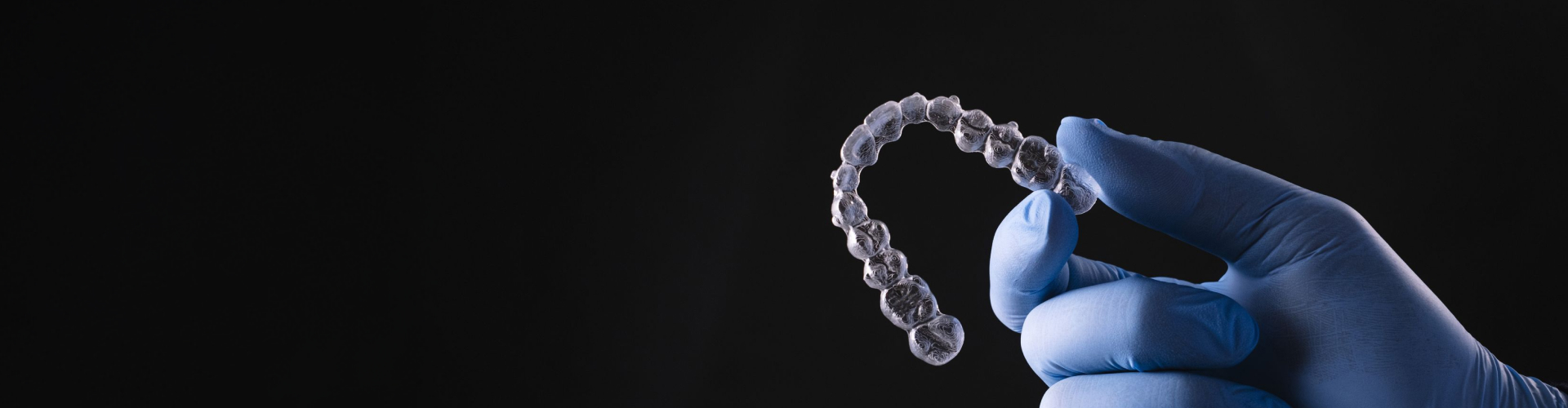 Dentist wearing blue gloves holding a clear Invisalign aligner tray against black background.